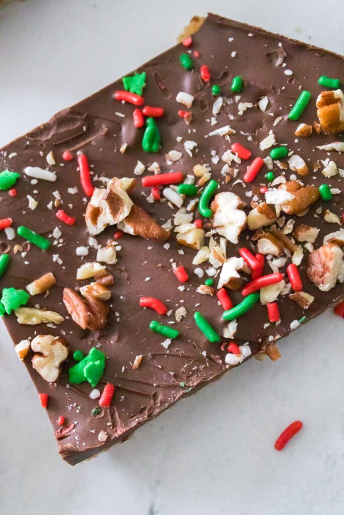 A rectangular piece of chocolate bark topped with red and green sprinkles, chopped nuts, and other small decorative toppings, resting on a white marble surface.