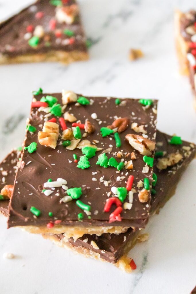 Two pieces of chocolate-covered toffee topped with chopped nuts and colorful red, green, and white sprinkles. They are stacked on a marble surface, offering a festive appearance.