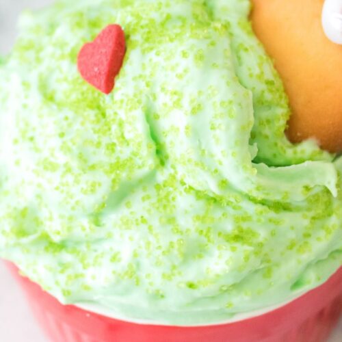 A red cup filled with green whipped cream topped with green sprinkles and a small red heart decoration. A hand is holding a vanilla wafer cookie, dipping it into the whipped cream. The background is a gray marble surface.
