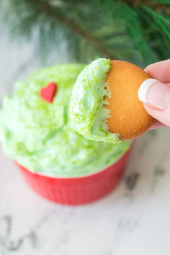A hand holds a cookie dipped in green frosting with a small red heart decoration. The frosting is in a red container on a marble surface, with pine branches in the background.