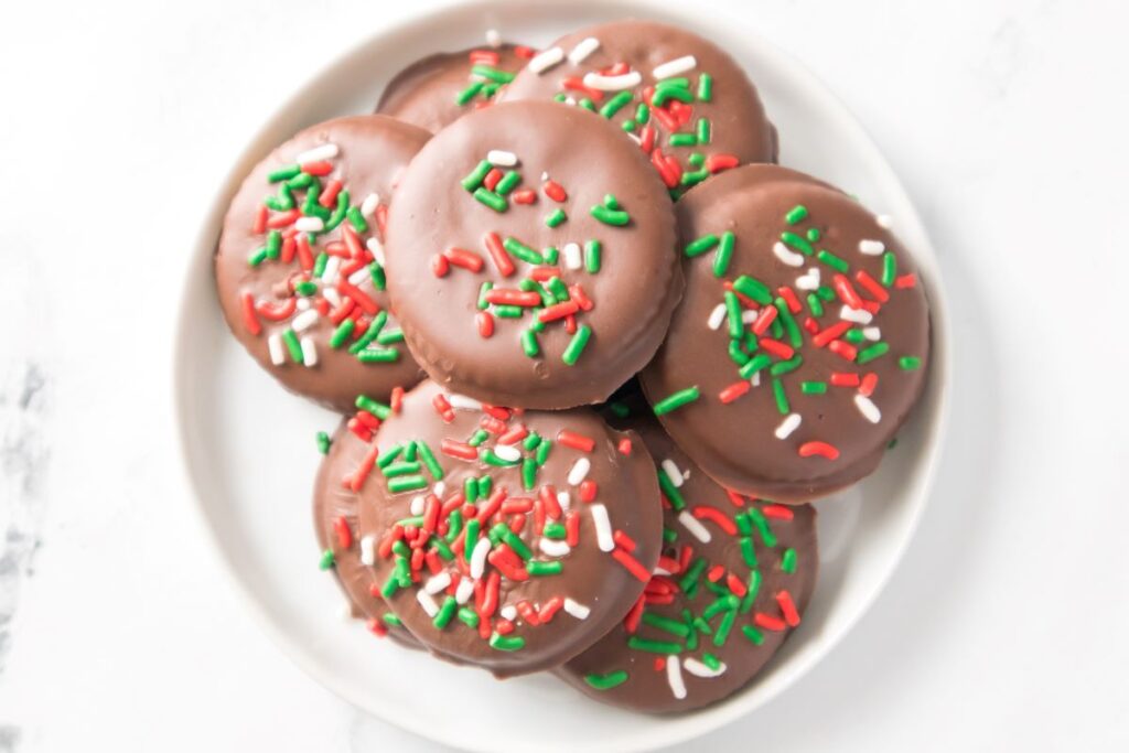 A plate of round chocolate-covered cookies topped with red, green, and white sprinkles. The cookies are neatly stacked and displayed on a white plate set against a light background.