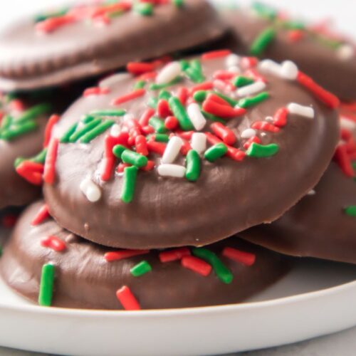 A plate of chocolate-covered cookies topped with red, green, and white sprinkles. The cookies are stacked in a pile, and the colorful sprinkles create a festive appearance.