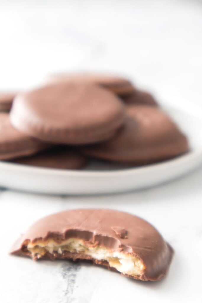 Close-up of a chocolate-covered cookie with a bite taken out, revealing a creamy filling. A plate in the background holds more cookies, slightly out of focus, on a white marble surface.