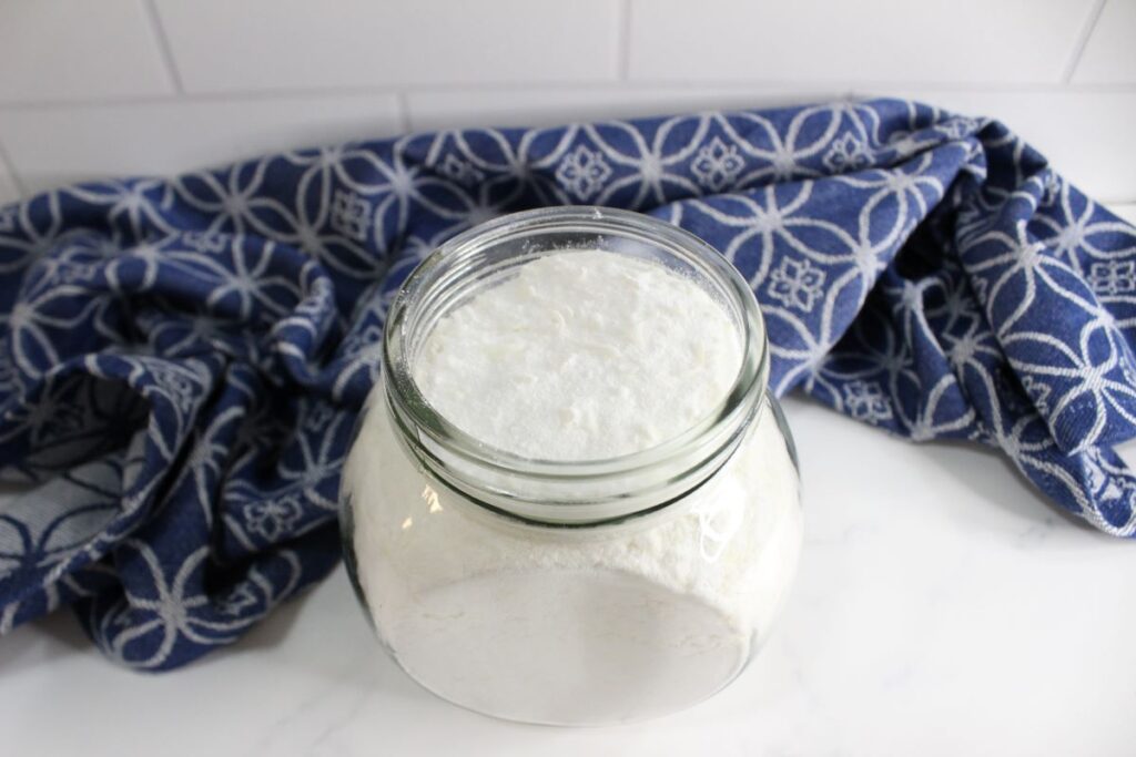 A glass jar filled with white powder, likely flour, sits on a white countertop. Behind the jar is a folded blue cloth with a white geometric pattern.