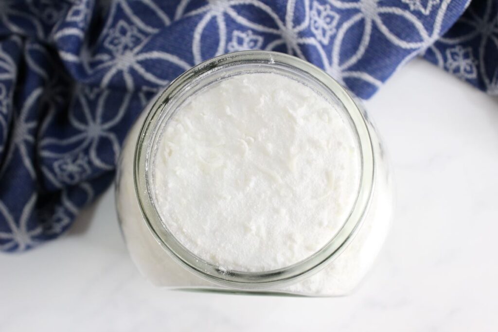 Top view of a glass jar filled with white, fluffy substance, likely a dairy product like yogurt or cream. A blue cloth with a white geometric pattern is partially visible in the background.