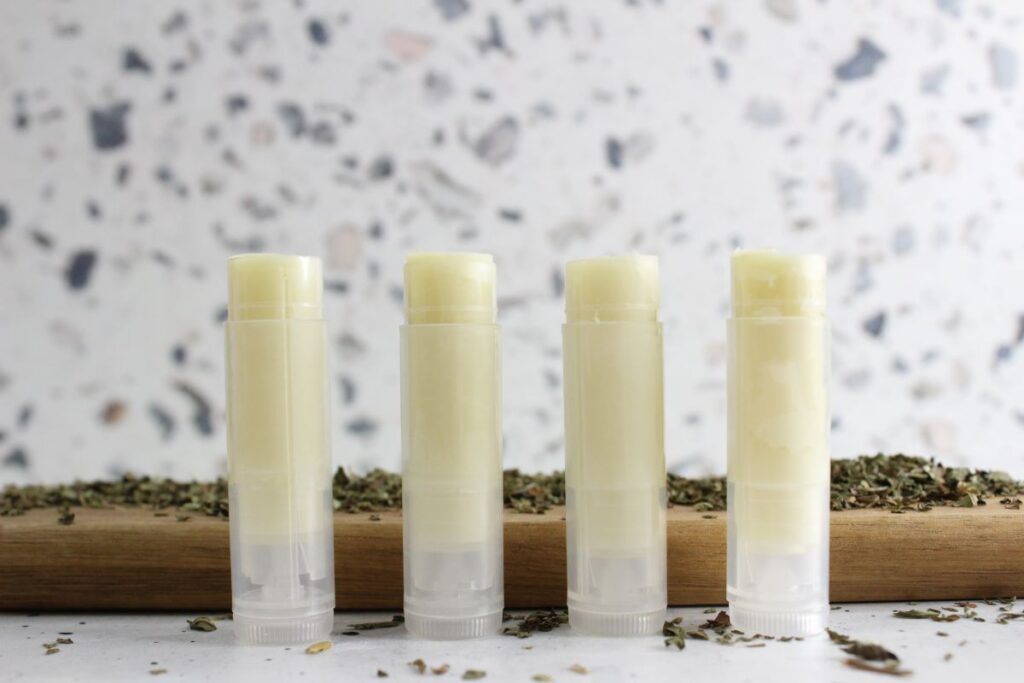 Four tubes of peppermint lip balm stand upright on a wooden surface, surrounded by dried herbs. The background features a speckled gray and white pattern.