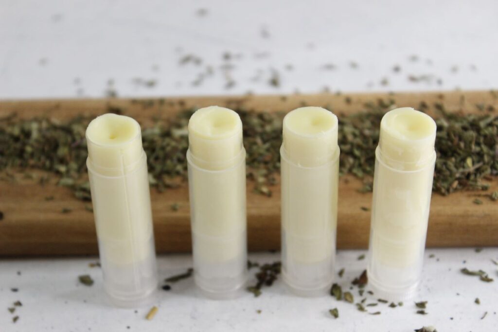 Four tubes of Peppermint Lip Balm stand upright on a wooden board, surrounded by scattered dried herbs. The background is a plain, light-colored surface, highlighting the soothing allure of minty freshness.