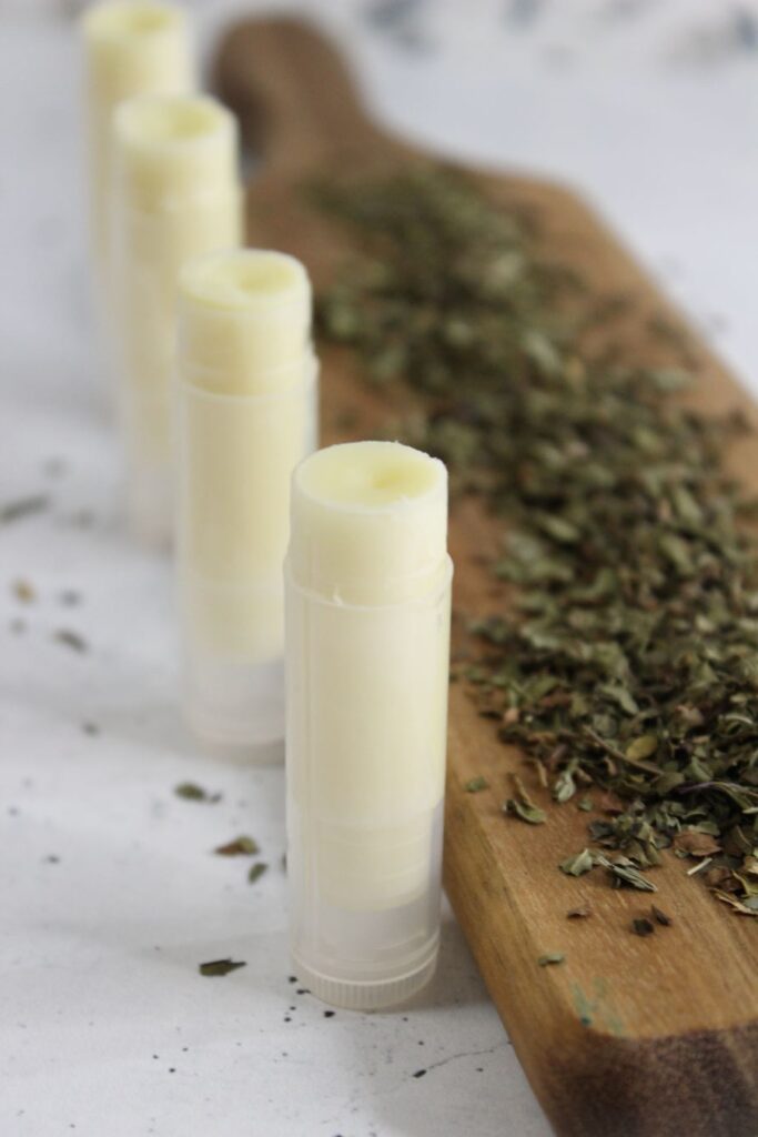 Several tubes of homemade peppermint lip balm are lined up on a white surface, next to a wooden board sprinkled with dried herbs.