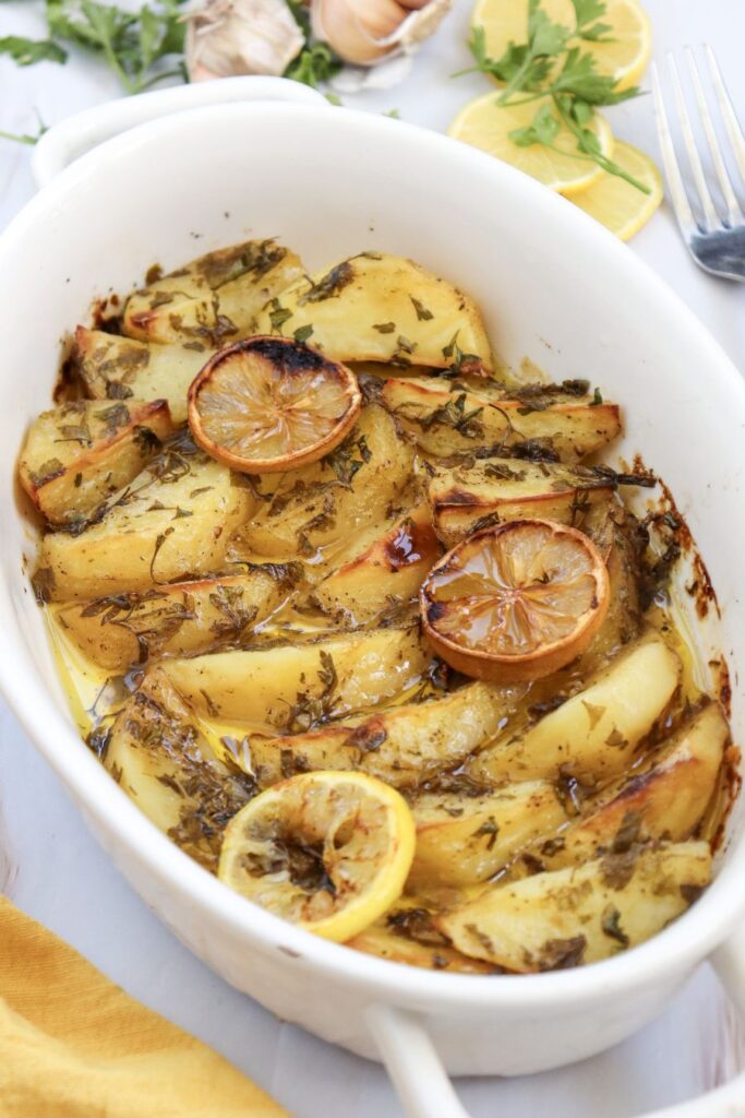 White baking dish with roasted potato wedges topped with lemon slices and herbs. The potatoes are golden brown with a slight shine from olive oil. Lemon slices and parsley garnish the dish, with a few lemon slices placed beside the dish.