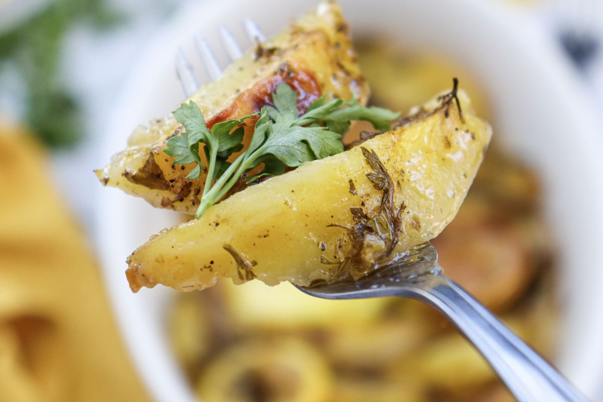 Close-up of a fork holding a seasoned potato wedge with a piece of arugula. The background shows a blurred dish filled with similar potato wedges, garnished with herbs and spices.