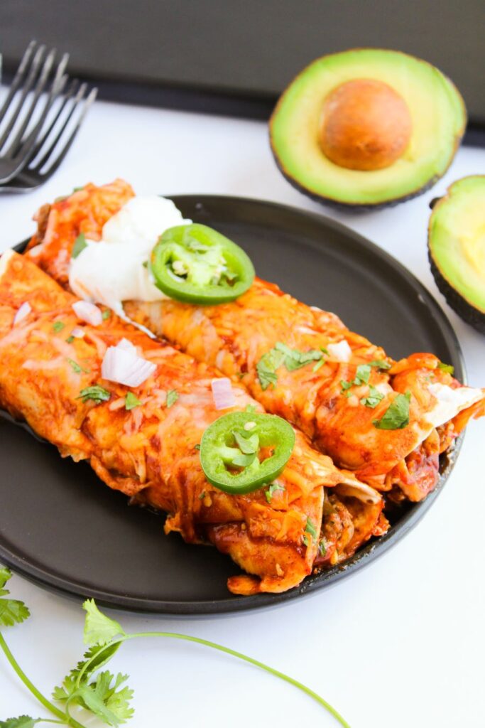 A plate of enchiladas topped with melted cheese, sour cream, sliced jalapeños, and cilantro. Avocado halves and forks are visible in the background.