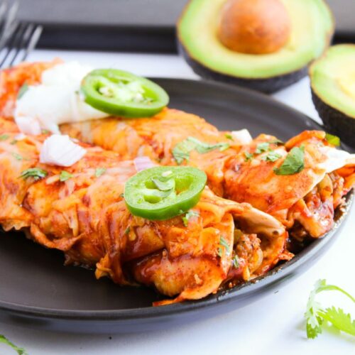 A black plate with two enchiladas topped with sliced jalapeños, sour cream, and onions. In the background, halved avocados and cilantro are visible. Four forks are placed on the side.