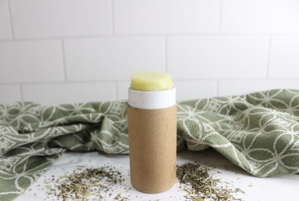 A tube of solid lotion stands upright on a white surface, surrounded by scattered herbs. The lotion is partially extended from the tube, which has a brown, eco-friendly appearance. A green patterned cloth is draped in the background.