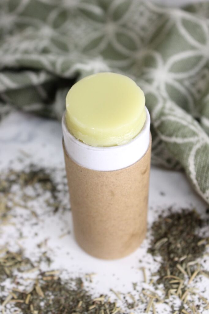 A natural deodorant stick with a cardboard casing is opened, showing the light green product. The stick is surrounded by scattered herbs, with a green cloth featuring a white circular pattern in the background.