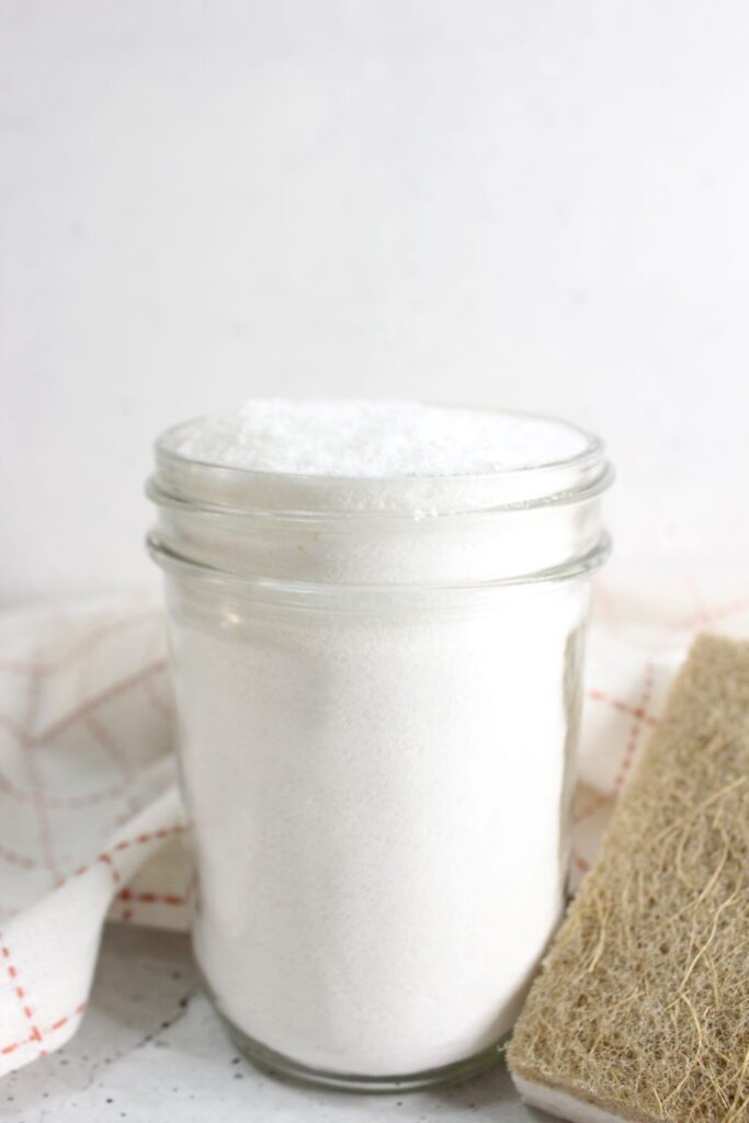 A glass jar filled with white powder, possibly sugar or salt, placed on a surface with a beige textured object nearby. A white cloth with red lines is partially visible in the background.