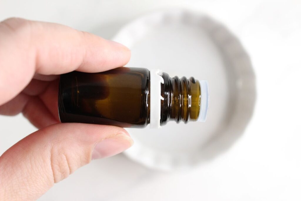 A hand holding a small, amber-colored glass bottle with a white cap, positioned above a round ceramic diffuser. The background is a soft, neutral surface.