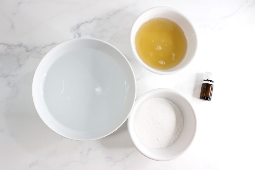 Top view of three white bowls on a marble surface. One bowl contains clear liquid, another has yellow liquid, and the third holds white powder. A small brown bottle with a white cap is next to the bowls.