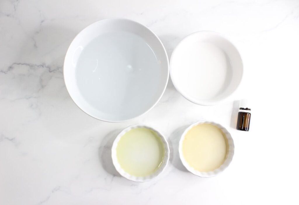 A flat lay of two bowls with clear and white liquids, a small brown bottle, and two ramekins with pale yellow and light green liquids, all on a white marble surface.