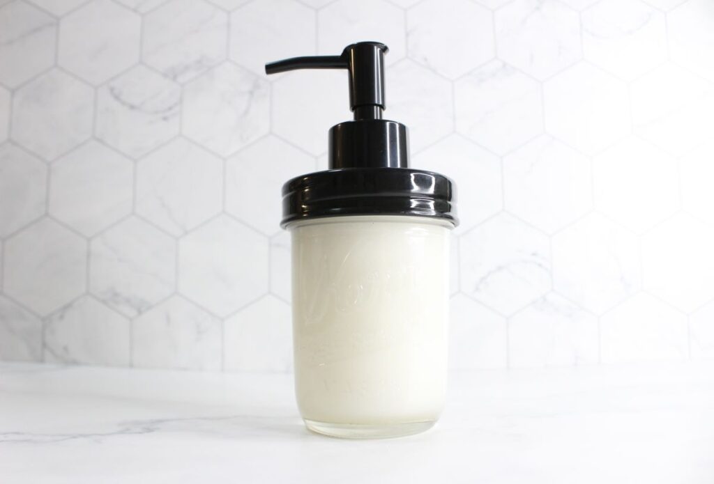 A pump dispenser filled with liquid is set against a white hexagonal tile background. The container appears to be a clear glass jar with a black pump lid.