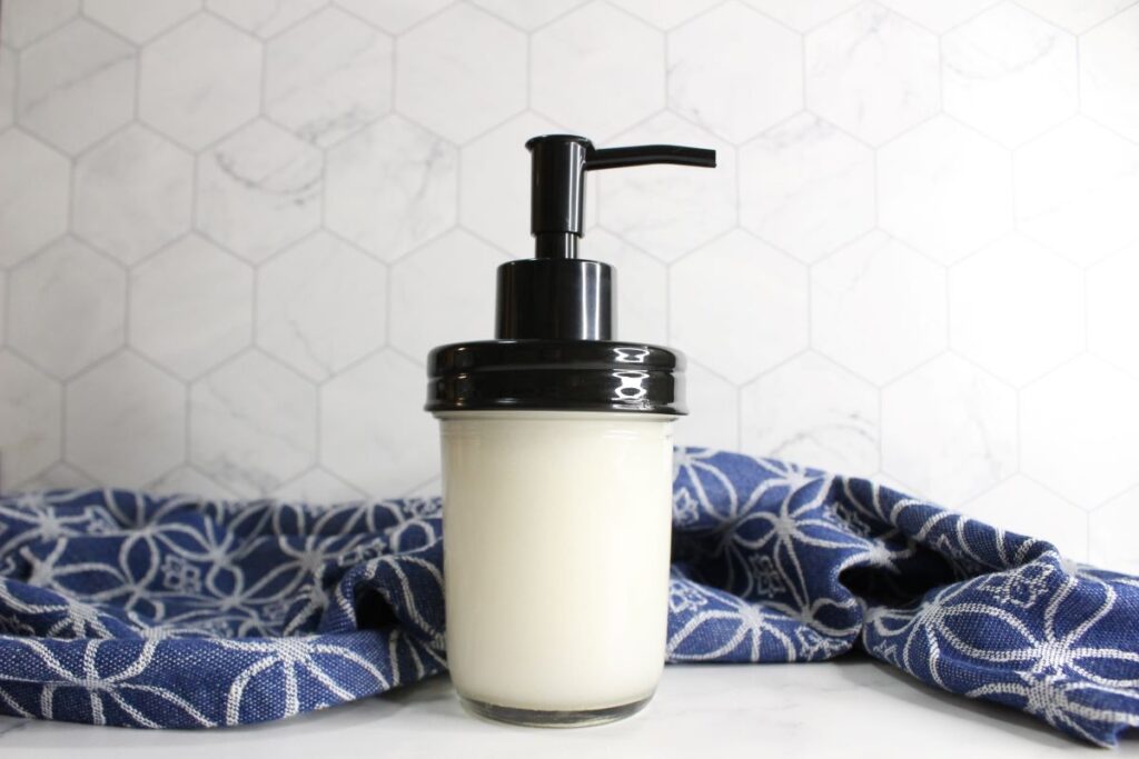 A glass jar with a pump top filled with white liquid sits on a white surface. It is set against a light hexagonal tile background, with a blue and white patterned cloth draped beside it.