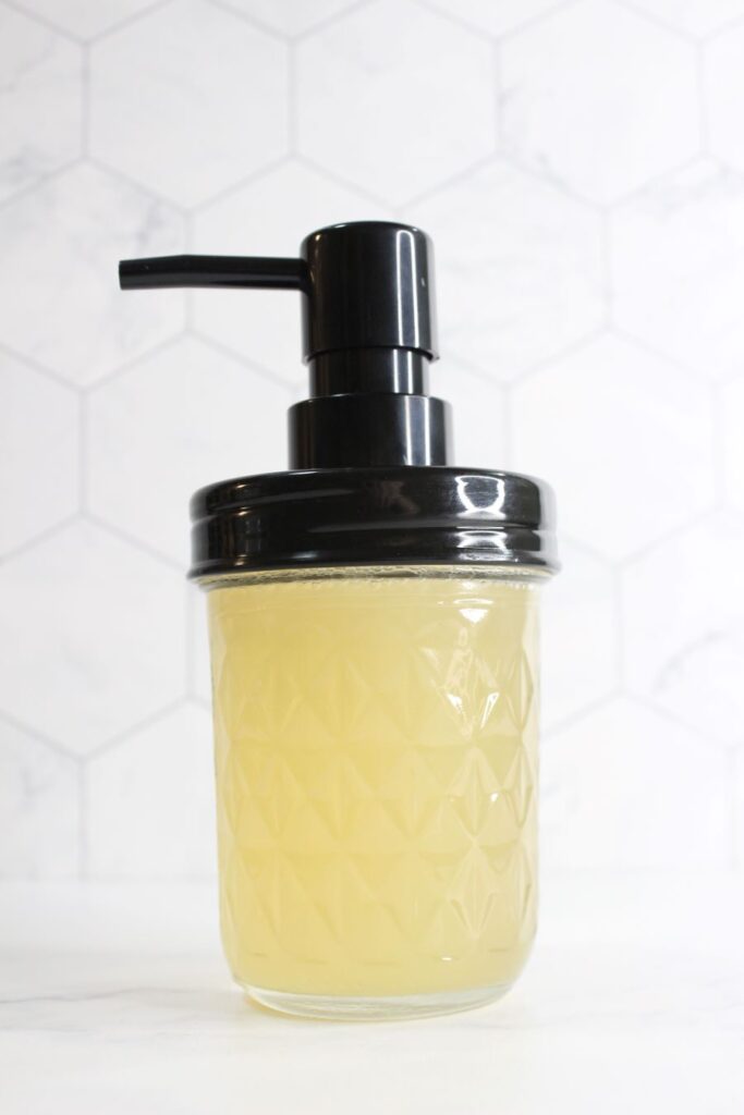 A clear, textured glass jar with a black pump lid filled with a light yellow liquid, set against a white hexagonal tile background.