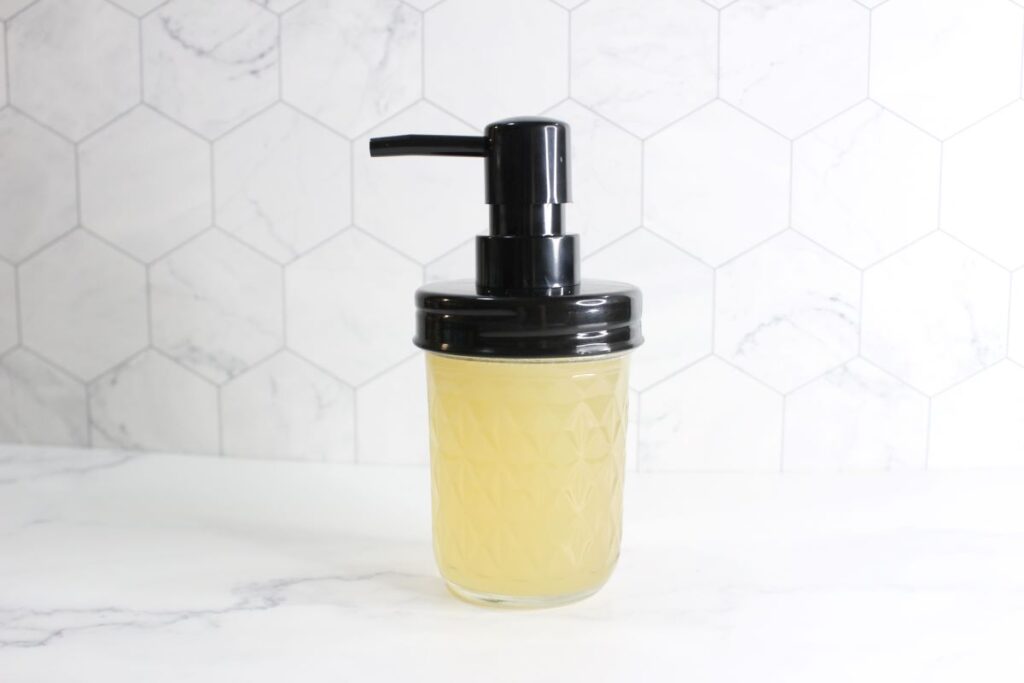 A clear glass jar with a black pump lid contains a yellow liquid inside. The jar sits on a white marble surface, and the background features a white hexagonal tile pattern.