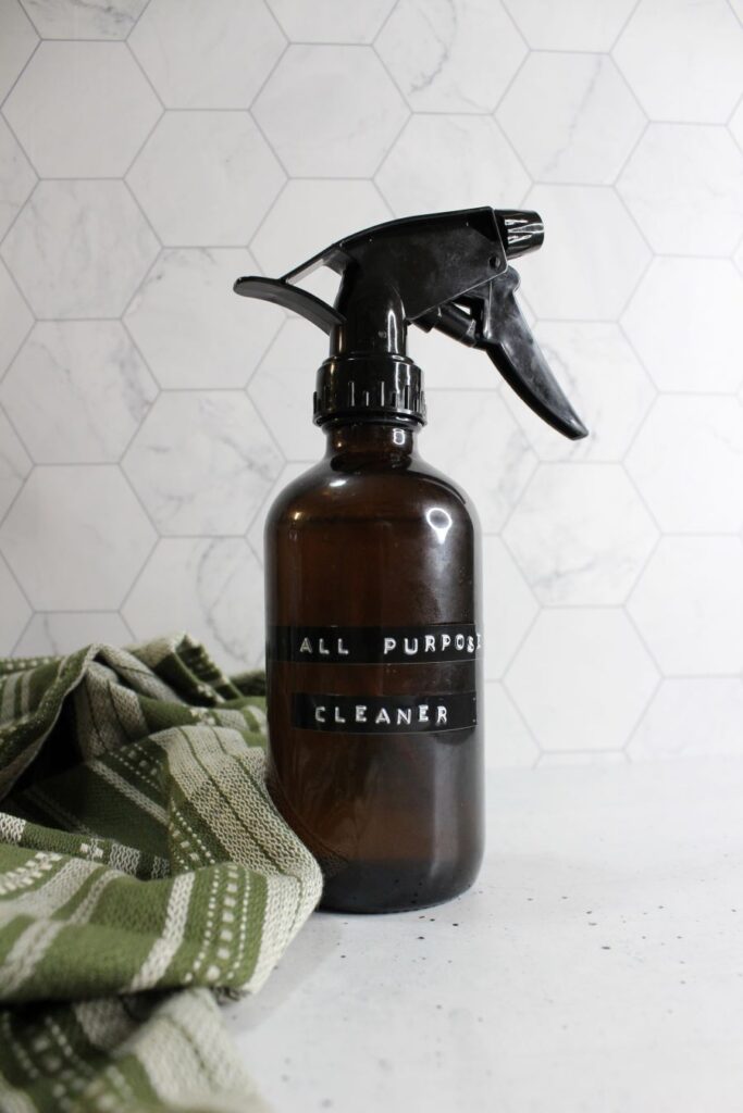 A brown spray bottle labeled "All Purpose Cleaner" stands on a light surface. A green patterned cloth is draped beside it. The background features white hexagonal tiles.