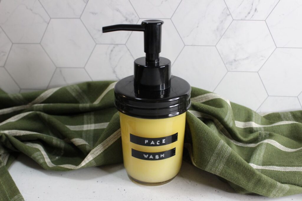 A bottle of face wash with a black pump top sits on a green cloth with white stripes, against a hexagonal tile background. The bottle has a label that reads "Face Wash" in capital letters.