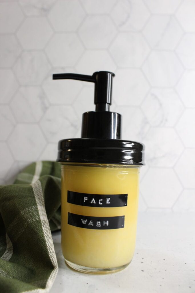A pump dispenser labeled "Face Wash" contains yellow liquid. It sits on a countertop with a hexagonal tile backsplash. A green and white cloth is draped nearby.