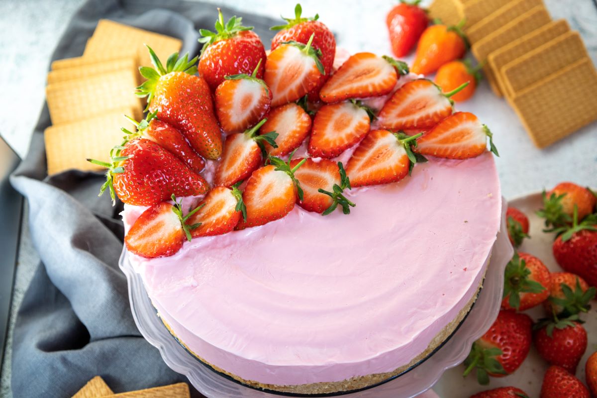 A pink strawberry cheesecake topped with fresh strawberries on a cake stand. Beside it are loose strawberries, square biscuits, and a gray cloth. The dessert is set on a light-colored surface.