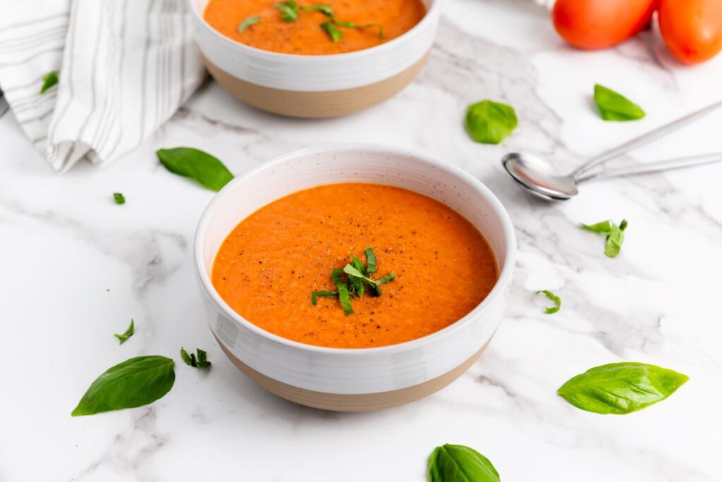 A bowl of creamy tomato soup garnished with fresh basil sits on a marble countertop. A second bowl and scattered basil leaves are in the background, along with a striped cloth and a spoon.