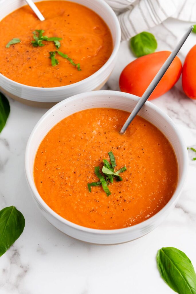 Two bowls of tomato soup garnished with chopped herbs are placed on a marble surface. Silver spoons rest in the bowls. Fresh basil leaves and ripe tomatoes are scattered around, adding a vibrant touch to the scene.