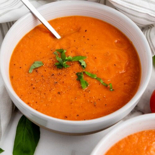 A bowl of creamy tomato soup garnished with freshly chopped basil and black pepper, with a spoon resting in it. The bowl is placed on a striped cloth next to a ripe tomato and basil leaves.