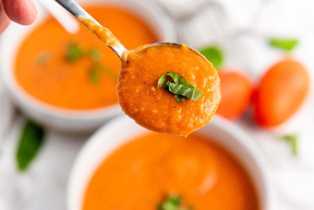 Close-up of a spoonful of creamy tomato soup garnished with basil. In the background, there are two bowls of the same soup and a couple of fresh tomatoes. The scene has a fresh and appetizing look.