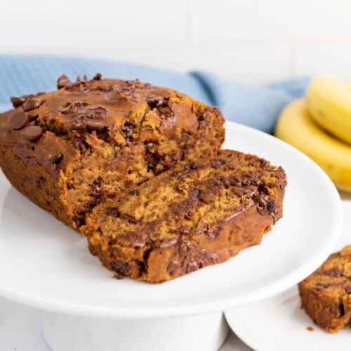 A slice of chocolate chip banana bread on a white cake stand, with scattered chocolate chips around. In the background, a bunch of bananas and a light blue cloth are visible.