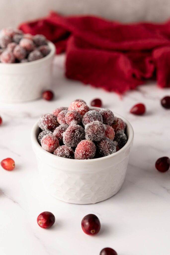 A white bowl filled with sugar-coated cranberries on a marble surface. A second bowl with more cranberries is in the background, along with scattered cranberries and a red cloth.