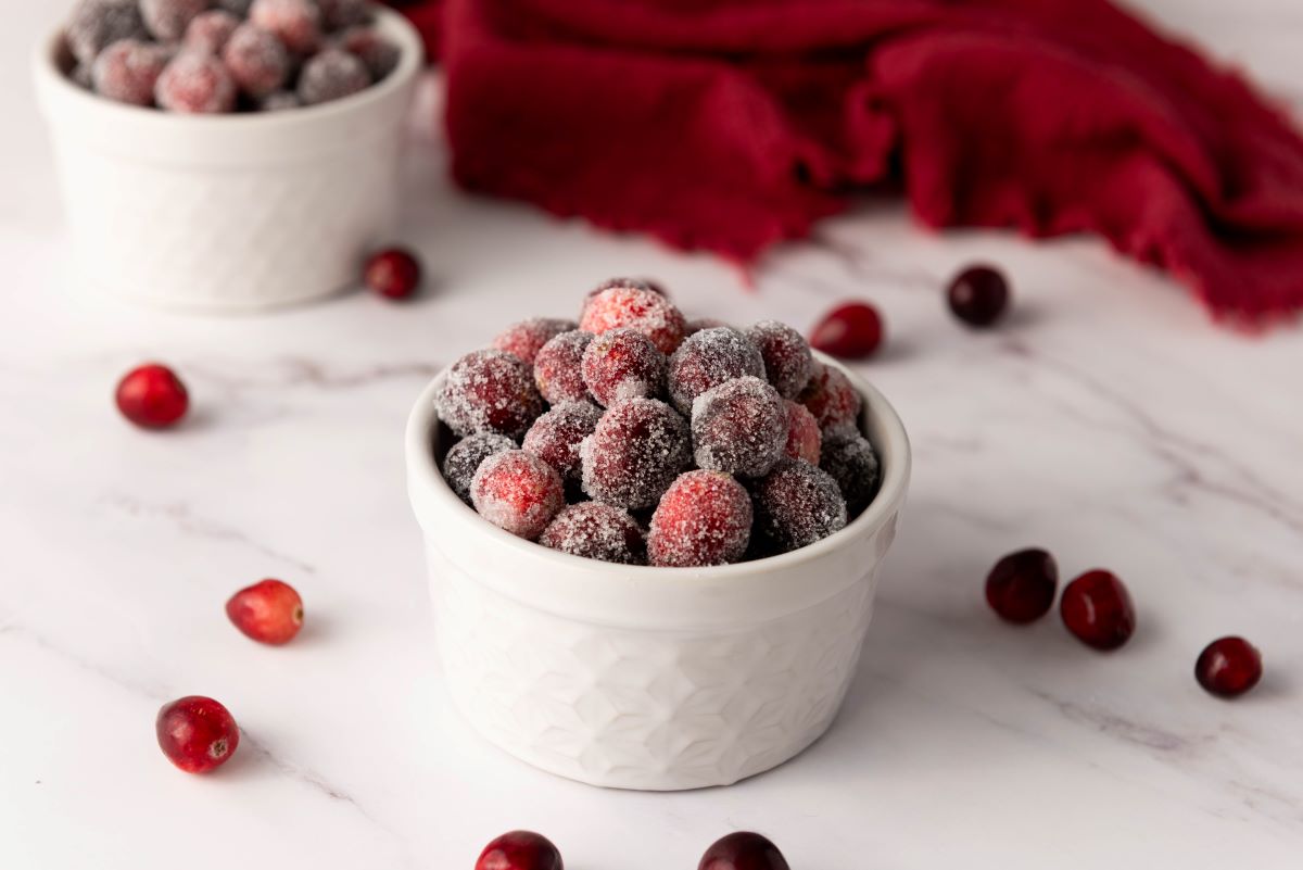 Two white bowls filled with sugared cranberries are placed on a marble surface. A few cranberries are scattered around, and a red cloth is partially visible in the background.