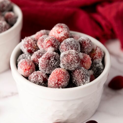 A white bowl filled with sugar-coated cranberries sits on a marble surface. A red cloth is in the background, with a few loose cranberries scattered around.