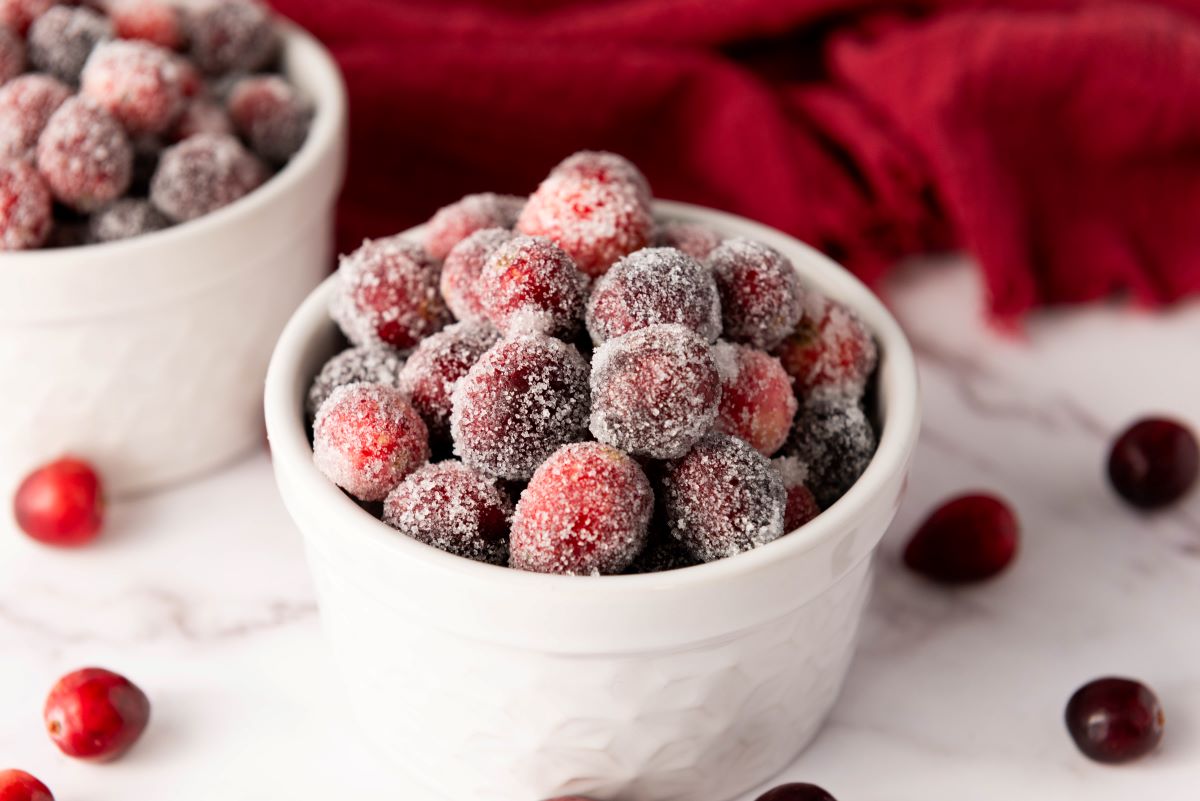 A white bowl filled with sugar-coated cranberries sits on a marble surface. A red cloth is in the background, with a few loose cranberries scattered around.