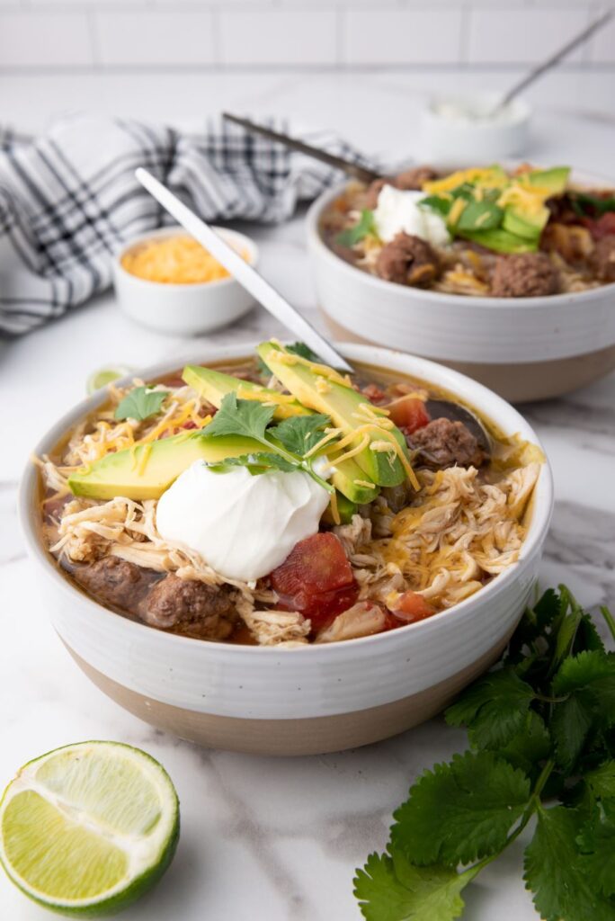 Two bowls of a hearty dish with shredded chicken, avocado slices, sour cream, and meatballs are served. Garnished with cilantro, with a halved lime and a bowl of shredded cheese nearby. A plaid cloth is in the background on a marble surface.