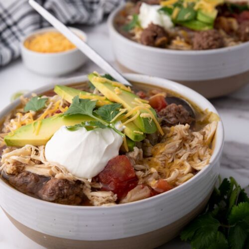 Bowls of chicken taco soup topped with sliced avocado, cheese, and sour cream on a marble surface. A striped towel, small dish of shredded cheese, and cilantro garnish in the background.