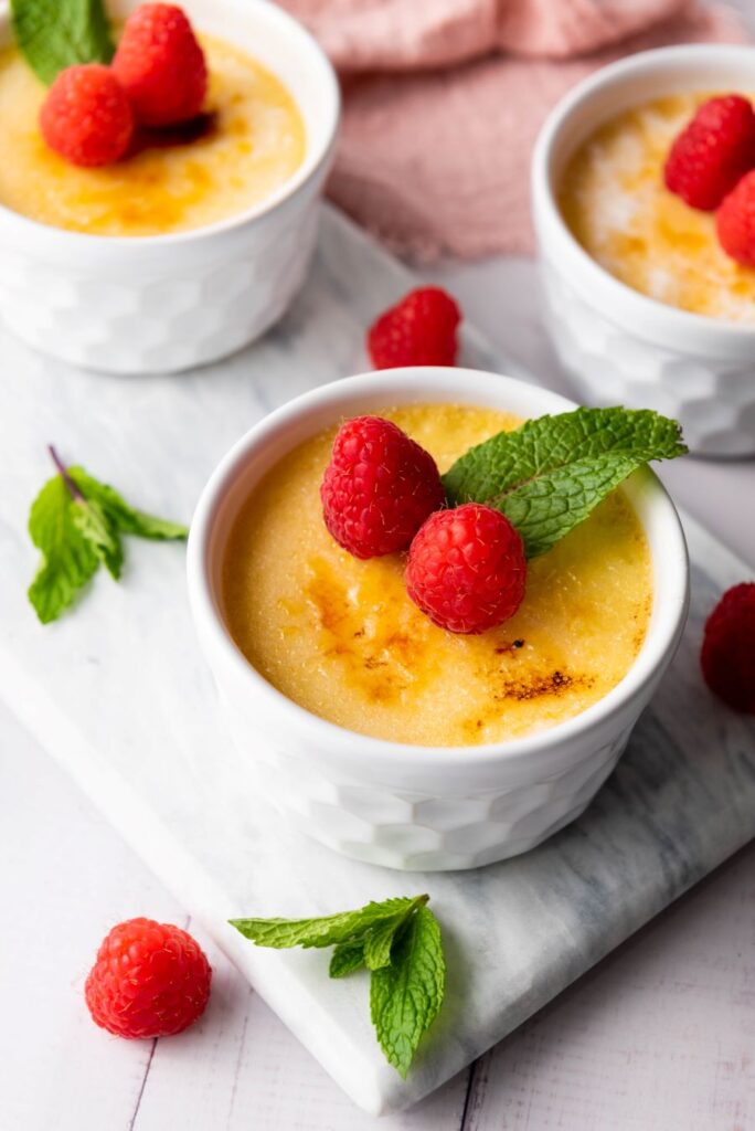 Baked custard in three white ramekins topped with fresh raspberries and mint leaves, placed on a marble surface. Additional raspberries and mint are scattered around for decoration.