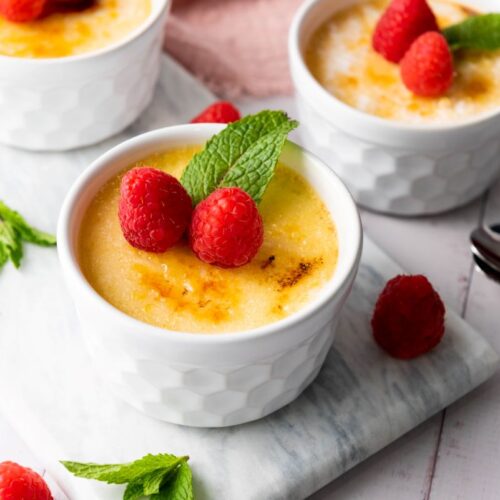 Three white ramekins filled with creamy crème brûlée, topped with fresh raspberries and mint leaves, placed on a marble board. Additional raspberries and mint leaves are scattered around, and two spoons rest nearby on a light surface.