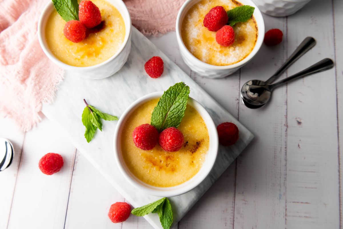 Three ramekins of crème brûlée topped with fresh raspberries and mint leaves are displayed on a white marble board. Additional raspberries and mint leaves are scattered around, with two spoons nearby on a wooden surface.