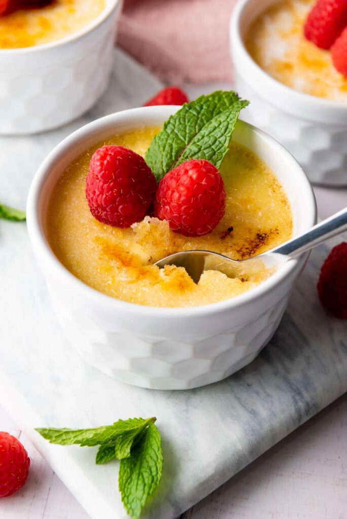 A dessert of crème brûlée in a white ramekin, topped with two raspberries and a mint sprig. A spoon is taking a portion, revealing the creamy texture underneath the caramelized sugar crust. More raspberries and mint leaves are scattered around.