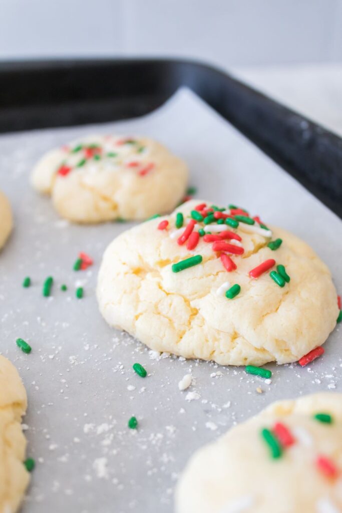 Close-up of freshly baked cookies on a baking sheet lined with parchment paper. The cookies are topped with red and green sprinkles, adding a festive touch. The background is blurred.