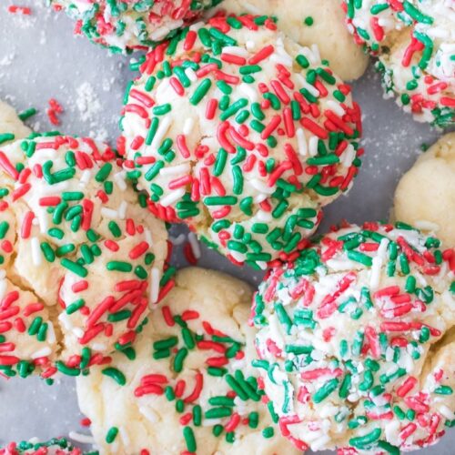 Festive cookies topped with red, green, and white sprinkles are scattered on a gray surface. Some cookies have more sprinkles, creating a textured, colorful pattern typical of holiday treats.