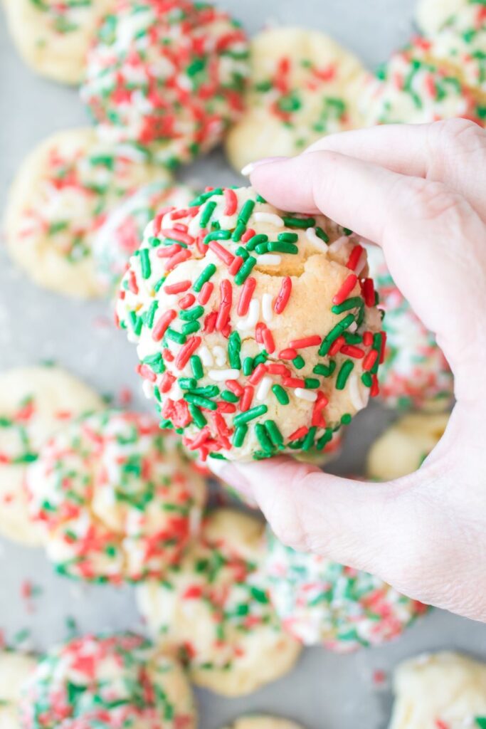 A hand holds a cookie topped with red and green sprinkles. More cookies with the same decoration are scattered on a surface below.