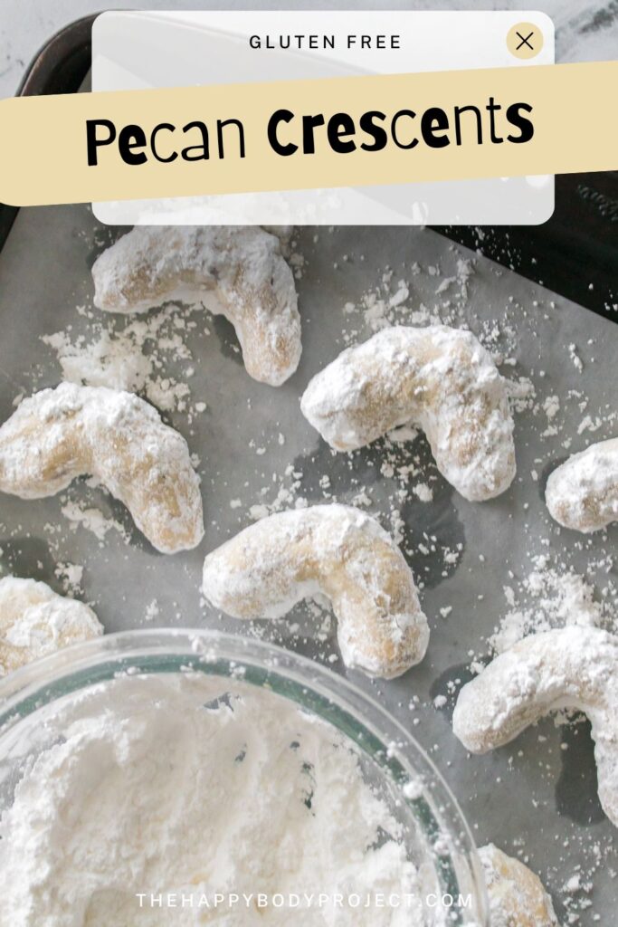 Gluten-free pecan crescents on a baking tray, dusted with powdered sugar. A mixing bowl with more sugar is partially visible. Text overlay reads "Pecan Crescents" and "thehappybodyproject.com.