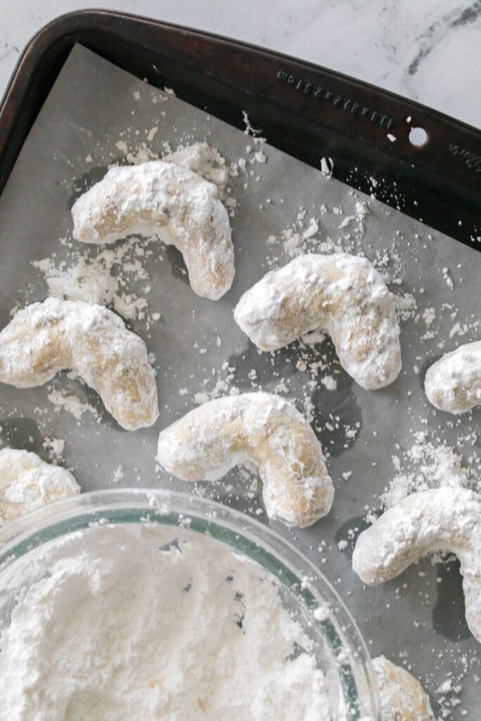 Powdered sugar-coated crescent-shaped cookies cool on a baking sheet lined with parchment paper. A bowl filled with powdered sugar is partially visible in the foreground.